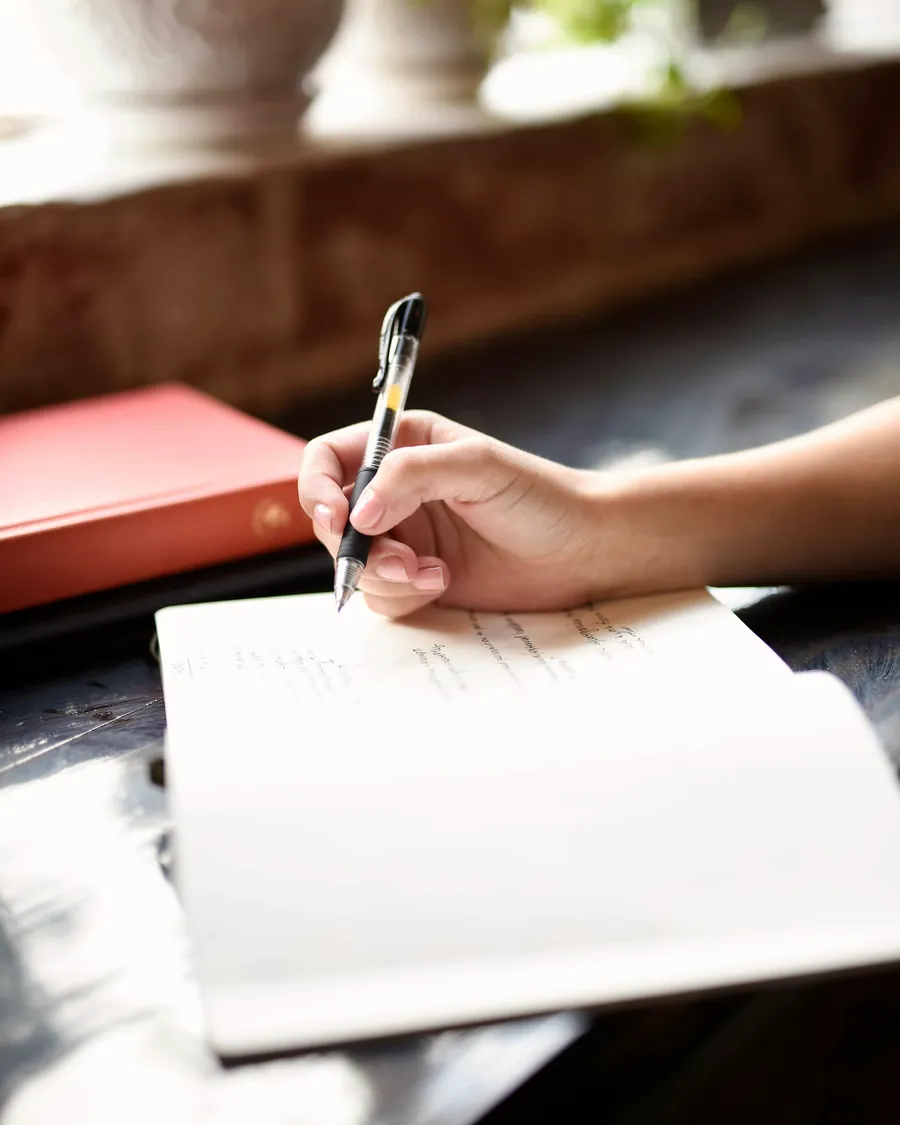Une main qui écrit à la plume sur une feuille, près d'une fenêtre, un livre rouge à côté