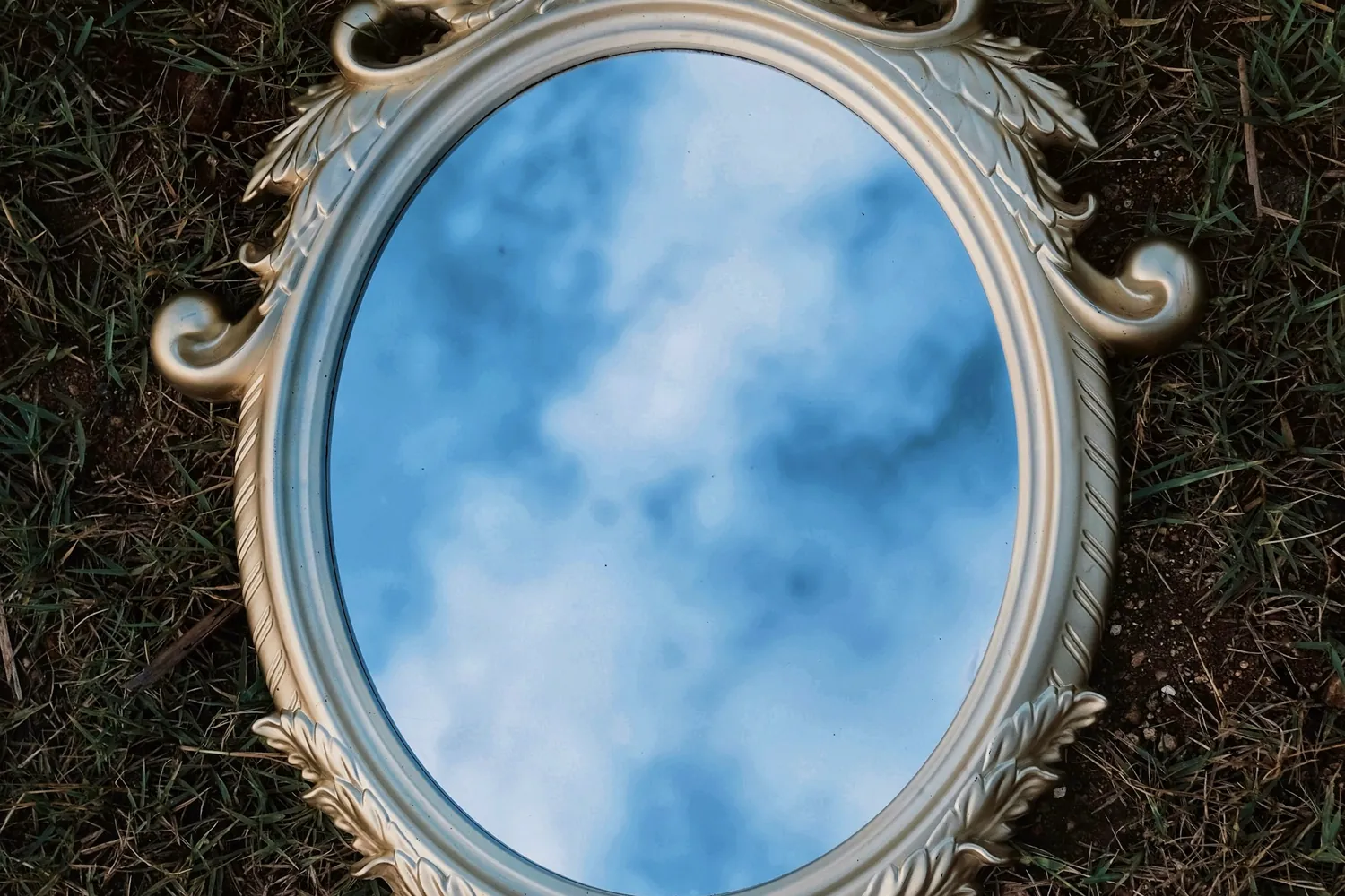 Un miroir ovale à cadre orné posé dans l'herbe et reflétant un ciel nuageux
